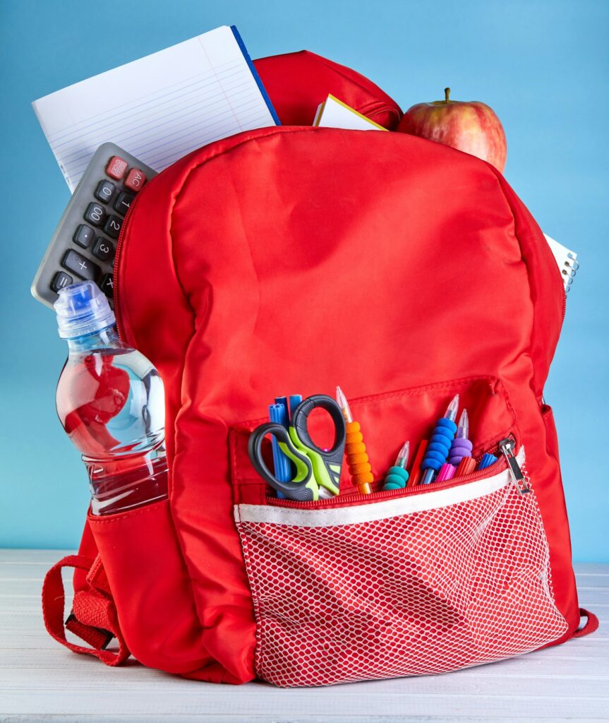 Stylish backpack with various school stationary on a white wooden table on a blue background